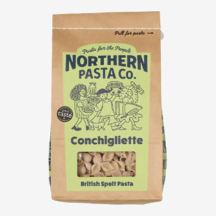 A clear plastic bag of small shell-shaped pasta shows tightly packed pale yellow conchigliette inside. A simple white label with blue and green accents names the brand and product, while nutrition facts and barcode appear on the back. The package sits on a plain surface. Soft shadows fall to the right.