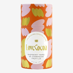 A pink and gold box sits open to reveal six glossy round truffles, each dusted with raspberry-red powder and speckled with glittering gold. The branded sleeve shows elegant script and delicate raspberry illustrations against a soft blush background.