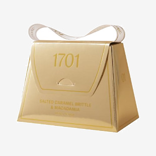 A handbag-shaped tin with a glossy caramel-brown exterior, embossed lettering and a tied ribbon. The lid opens to reveal golden salted caramel brittle and pale macadamia honey nougat pieces studded with glossy nuts, set against a crinkled paper liner.